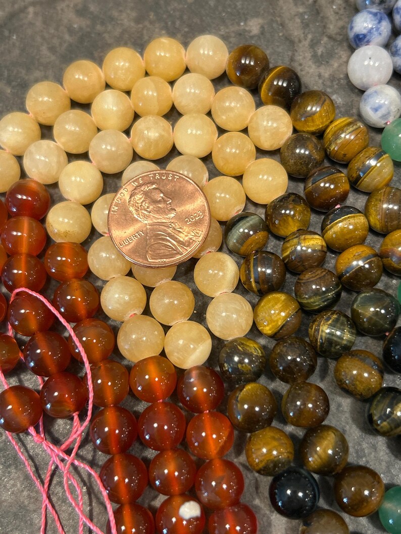 May include: A close-up of three strands of gemstone beads. The top strand is made of light yellow beads, the middle strand is made of brown and gold tiger's eye beads, and the bottom strand is made of reddish-brown carnelian beads. A US penny is included for scale.