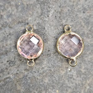 May include: Two round, faceted, light pink gemstone connectors with gold-colored metal settings. Each connector has a loop at the top and bottom for attaching to other jewelry components. The gemstones have a geometric cut, reflecting light.