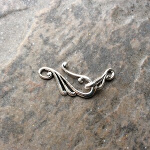 May include: A silver-toned clasp with an ornate design, featuring curved lines and a hook closure. The clasp has a decorative, stylized wing-like shape on each end, suitable for jewelry or craft projects.
