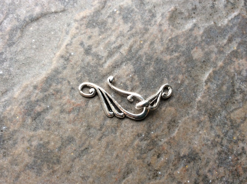 May include: A silver-toned clasp with an ornate design, featuring curved lines and a hook closure. The clasp has a decorative, stylized wing-like shape on each end, suitable for jewelry or craft projects.