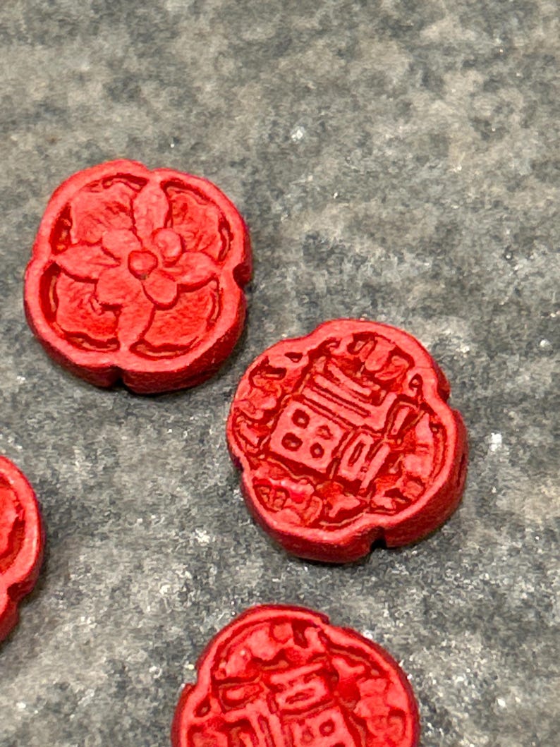 Pu&ograve; includere: Primo piano di diversi piccoli oggetti circolari rosso vivo. Ogni oggetto presenta un design in rilievo dettagliato, con motivi floreali e geometrici. Gli oggetti sono sparsi su una superficie grigia strutturata.