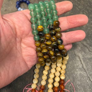 May include: A set of six strands of gemstone beads. The beads are round and smooth. The top strand is green, the second is brown and gold, the third is cream, the fourth is orange, and the bottom two are red.