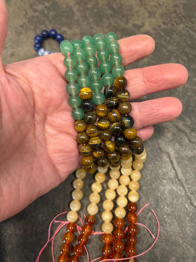 May include: A set of six strands of gemstone beads. The beads are round and smooth. The top strand is green, the second is brown and gold, the third is cream, the fourth is orange, and the bottom two are red.