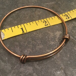 May include: A copper expandable bangle bracelet measuring 2 inches in diameter. The bracelet has a simple design with two small knots on each end.