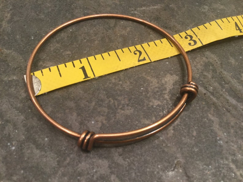 May include: A copper expandable bangle bracelet measuring 2 inches in diameter. The bracelet has a simple design with two small knots on each end.