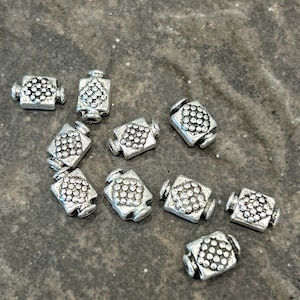 May include: A collection of small, silver-colored metal beads. Each bead is rectangular with a textured, dotted design on the front and back. The beads are scattered on a textured, gray surface.