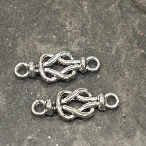 May include: Two silver metal clasps with a knot design. The clasps have two loops on each end.