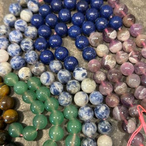 May include: A close-up of a variety of gemstone beads, including green, blue, and purple. The beads are arranged in a circular pattern, with the green beads in the center and the blue and purple beads surrounding them.