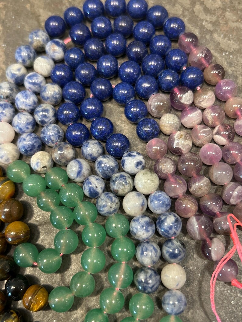 May include: A close-up of a variety of gemstone beads, including green, blue, and purple. The beads are arranged in a circular pattern, with the green beads in the center and the blue and purple beads surrounding them.