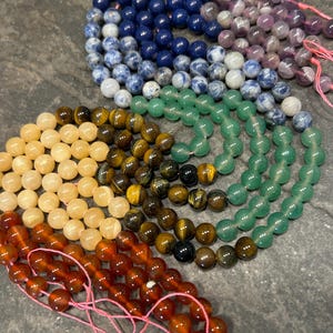 May include: A set of seven strands of gemstone beads, each strand a different color. The colors are blue, purple, green, yellow, orange, red, and brown. The beads are strung on pink thread.