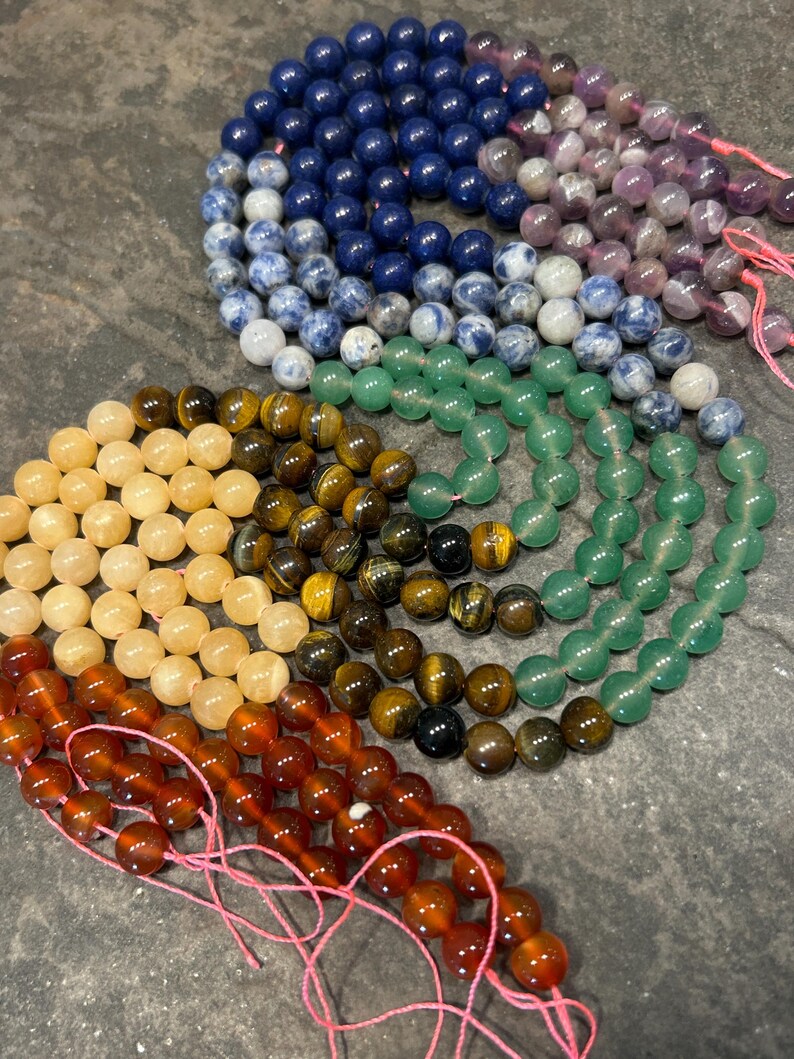 May include: A set of seven strands of gemstone beads, each strand a different color. The colors are blue, purple, green, yellow, orange, red, and brown. The beads are strung on pink thread.