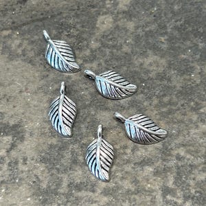May include: Five silver-colored leaf charms with detailed veining. Each charm has a small loop at the top for attaching to jewelry or other crafts. The charms are scattered on a textured gray surface.
