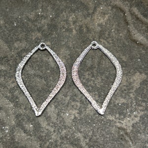 May include: Two silver, teardrop-shaped metal earring blanks with a textured surface.