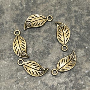 May include: Five antique gold-colored leaf charms arranged in a circle. Each charm has a detailed leaf vein design and a small loop at the top for attaching to jewelry or other crafts. The charms are on a textured gray surface.