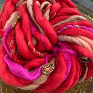 May include: A large skein of yarn in shades of red, pink, and beige with gold accents. The yarn is arranged in a loose coil and features decorative embellishments.