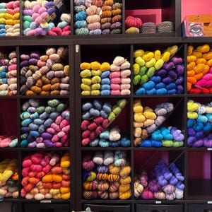 May include: A display of colorful yarn skeins arranged in a black shelving unit. The yarn is organized by color, with shades of pink, yellow, blue, purple, and orange. The yarn is neatly stacked, creating a visually appealing and organized craft supply display.