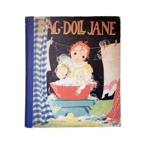 May include: Vintage children's book cover titled "Rag-Doll Jane." The cover features an illustration of a rag doll in a red basin filled with bubbles. The doll is washing another doll. The background is dark with bubbles and laundry hanging on a clothesline.