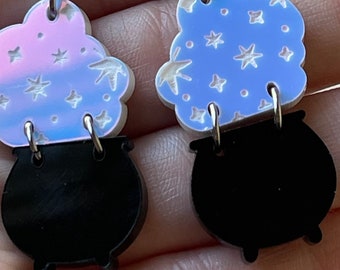 May include: A pair of dangle earrings featuring a cloud-shaped top with star and leaf cutouts, in iridescent pink and blue hues, connected to a black cauldron. The earrings have silver-colored ear wires.