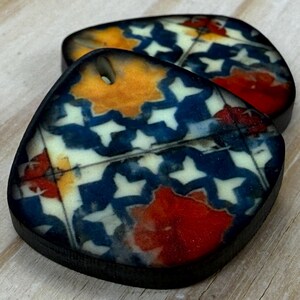 May include: Two decorative, teardrop-shaped pendants with a geometric pattern. The design features white star shapes on a navy blue background, with orange and red accents. Each pendant has a small hole at the top and a black border.