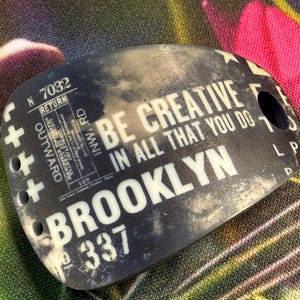 May include: A black and white tag with the words "BE CREATIVE IN ALL THAT YOU DO BROOKLYN" printed on it. The tag has a hole and several other printed details, including the number "337".