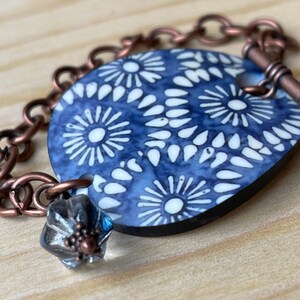 May include: A blue and white patterned ceramic bracelet with a copper chain and a toggle clasp. The bracelet has a small, clear glass bead with a silver accent.
