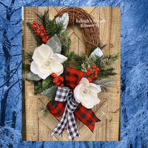 May include: A rustic wreath with a grapevine base, adorned with white magnolia flowers, red berries, and evergreen sprigs. A red and black buffalo plaid bow and a black and white checkered bow add a festive touch. The text "Kaleigh's Wreath & Decor Co." is visible.