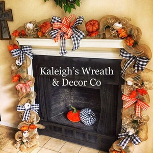 May include: A fireplace mantel decorated with a burlap garland, orange pumpkins, and black and white checkered bows. The garland is adorned with faux fall foliage and small white pumpkins. The text "Kaleigh's Wreath & Decor Co" is visible.