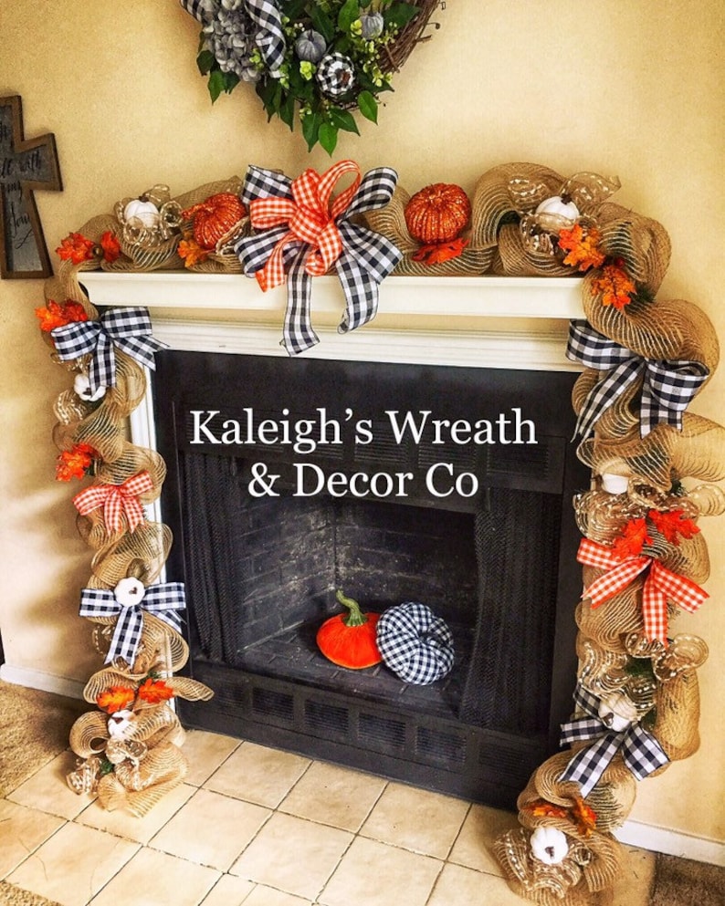 May include: A fireplace mantel decorated with a burlap garland, orange pumpkins, and black and white checkered bows. The garland is adorned with faux fall foliage and small white pumpkins. The text "Kaleigh's Wreath & Decor Co" is visible.