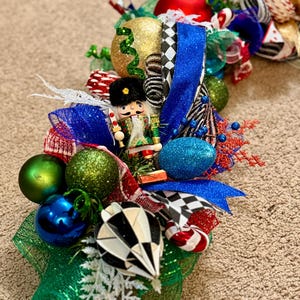 May include: A festive Christmas wreath with a variety of ornaments, including gold, green, and blue spheres, and a decorative nutcracker figure. The wreath also features blue and red ribbons, and a black and white checkered pattern.