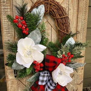 May include: A rustic grapevine wreath adorned with white magnolia flowers, red berries, and evergreen sprigs. Accented with a red and black buffalo plaid bow and a black and white checkered bow, creating a festive holiday decoration.