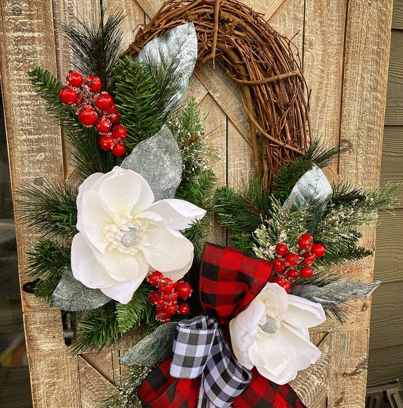 May include: A rustic grapevine wreath adorned with white magnolia flowers, red berries, and evergreen sprigs. Accented with a red and black buffalo plaid bow and a black and white checkered bow, creating a festive holiday decoration.