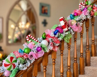 May include: A festive Christmas garland draped over a wooden staircase railing, featuring flocked pine branches, red and white candy cane ornaments, round peppermint-style decorations, pink mesh ribbon, and colorful shiny baubles in green, blue, and pink.