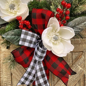 May include: A decorative wreath featuring large white magnolia flowers, red berries, and green pine needles. The wreath is accented with a buffalo plaid bow in red and black, and a black and white checkered bow. The background is a rustic wooden door.