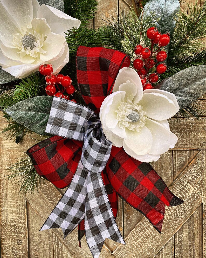 May include: A decorative wreath featuring large white magnolia flowers, red berries, and green pine needles. The wreath is accented with a buffalo plaid bow in red and black, and a black and white checkered bow. The background is a rustic wooden door.