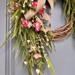 K&ouml;nnte beinhalten: Ein Weinrebenkranz, der mit rosa Bl&uuml;ten, Gr&uuml;n und einem braunen Juteband verziert ist. Der Kranz h&auml;ngt an einer grauen T&uuml;r.