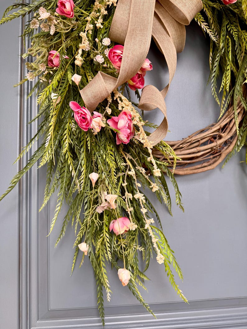 K&ouml;nnte beinhalten: Ein Weinrebenkranz, der mit rosa Bl&uuml;ten, Gr&uuml;n und einem braunen Juteband verziert ist. Der Kranz h&auml;ngt an einer grauen T&uuml;r.