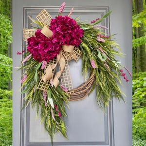 May include: A pink and green wreath with a burlap bow and a large pink flower cluster. The wreath is hanging on a gray door.