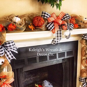 May include: A fall-themed mantel garland featuring burlap mesh, pumpkins, and fall leaves. The garland is adorned with black and white checkered bows and orange accents. The text "Kaleigh's Wreath & Decor Co." is visible.