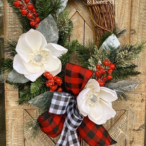 May include: A decorative wreath featuring a grapevine base adorned with white magnolia flowers, red berries, and evergreen sprigs. A red and black buffalo plaid bow and a black and white checkered bow add a festive touch. The text "Kaleigh's Wreath & Decor Co." is visible.