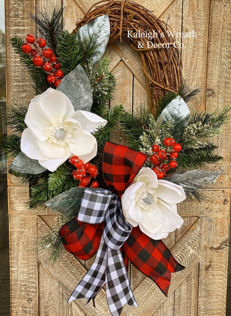 May include: A decorative wreath featuring a grapevine base adorned with white magnolia flowers, red berries, and evergreen sprigs. A red and black buffalo plaid bow and a black and white checkered bow add a festive touch. The text "Kaleigh's Wreath & Decor Co." is visible.