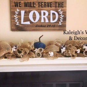 K&ouml;nnte beinhalten: Ein rechteckiges Holzschild mit dem Text WE WILL SERVE THE LORD Joshua 24:15 &uuml;ber einem Kaminsims, der mit einer Jute-Girlande, wei&szlig;en Baumwollbl&uuml;ten, kleinen Lichterketten und einem einzelnen dunkelblauen Zierk&uuml;rbis geschm&uuml;ckt ist.