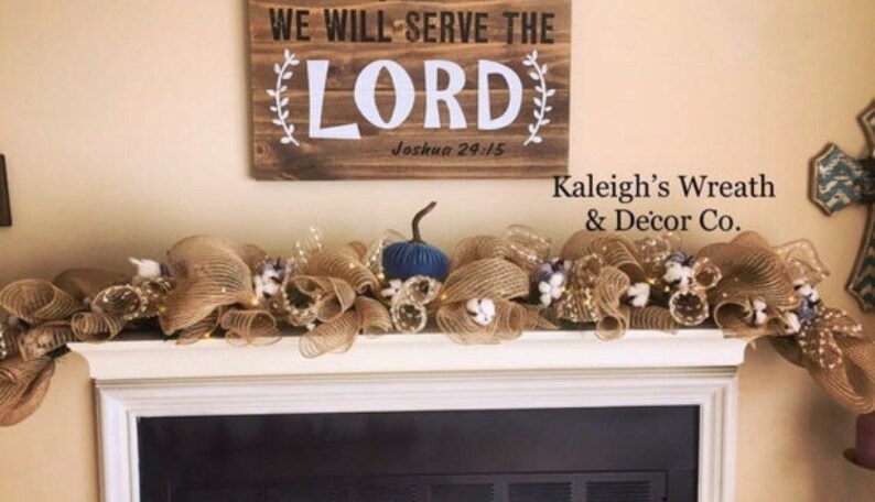 K&ouml;nnte beinhalten: Ein rechteckiges Holzschild mit dem Text WE WILL SERVE THE LORD Joshua 24:15 &uuml;ber einem Kaminsims, der mit einer Jute-Girlande, wei&szlig;en Baumwollbl&uuml;ten, kleinen Lichterketten und einem einzelnen dunkelblauen Zierk&uuml;rbis geschm&uuml;ckt ist.