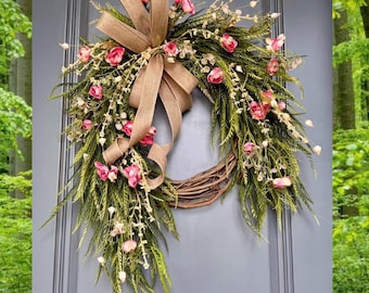 May include: A wreath made of green branches and pink flowers with a burlap bow. The wreath is hanging on a gray door.