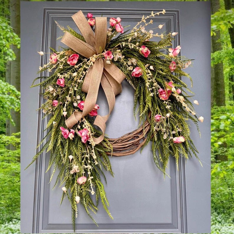 K&ouml;nnte beinhalten: Ein Kranz aus gr&uuml;nen Zweigen und rosa Bl&uuml;ten mit einer Schleife aus Sackleinen. Der Kranz h&auml;ngt an einer grauen T&uuml;r.