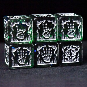 May include: Six green and black dice with white skeletal hand designs. The dice are arranged in two rows of three, with the hand symbols etched into the faces. The dice have a translucent quality, with hints of blue and green.