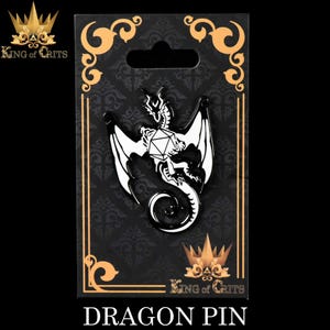 May include: A black and white enamel pin featuring a dragon curled around a white twenty-sided die. The pin is on a black background with gold trim and the text "King of Crits" in gold letters. The text "Dragon Pin" is below the image.