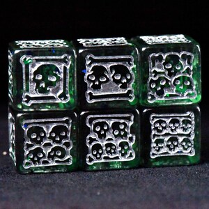 May include: Six green dice with silver skull and crossbones designs. Each die face features a different arrangement of skulls, creating a unique and thematic set. The dice are likely used for tabletop games.