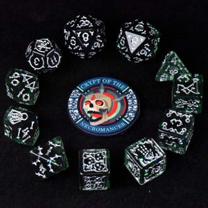 May include: A collection of dice and a circular patch with a skull design. The dice are black and green with white markings, including skulls and bones. The patch reads "Crypt of the Necromancer" with a skull in the center.