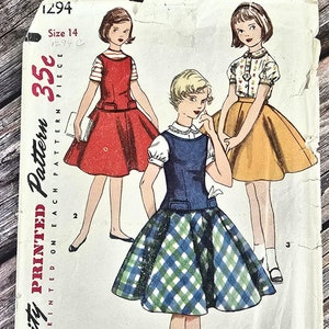 May include: Vintage Simplicity sewing pattern for girls' dresses. The pattern is for sizes 14 and includes three different styles: a red dress with a white collar, a blue and green plaid dress with a white collar, and a yellow dress with a white floral print.