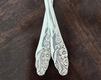 May include: Two silver spoons with ornate floral designs on the handles. The spoons are crossed over each other.